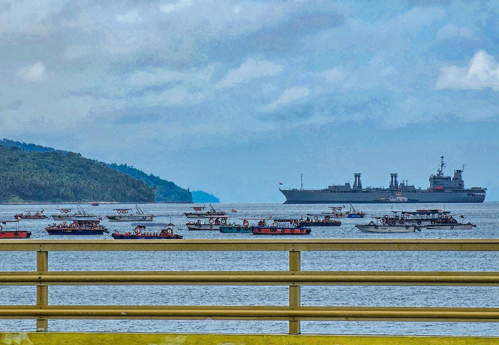 Multiple passenger ferries and an Indian naval ship anchored near the forested coastline of Andaman Islands, viewed from a bridge